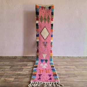 May include: A long, rectangular runner rug featuring a vibrant pink central field decorated with various geometric shapes in white, green, orange, and blue. The edges are bordered by a colourful, multi-patterned stripe design with fringe at the end.
