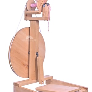 Puede incluir: Una rueca de madera con una gran rueda circular y un huso más pequeño para enrollar el hilo. La rueda es de madera clara y tiene un pedal. Se ve hilo rosa en el huso.