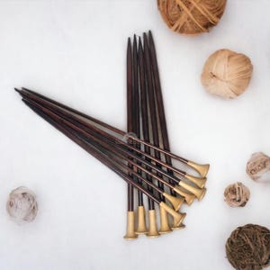 May include: Dark brown knitting needles with gold-coloured tips are arranged with balls of yarn in shades of brown, beige, and burgundy on a white background. The yarn balls vary in size and texture.