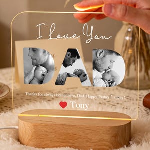Könnte beinhalten: Eine personalisierte Acryl-Fotoanzeige mit dem Wort "DAD" und dem Satz "I Love You". Die Anzeige zeigt drei Schwarz-Weiß-Fotos und einen Holzsockel. Der Name "Tony" und ein Herz befinden sich unten.