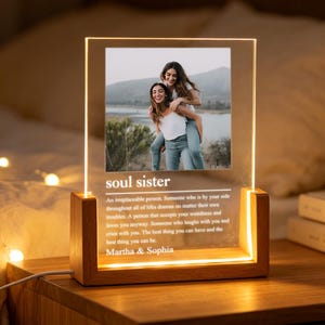 Könnte beinhalten: Eine rechteckige Acryl-Fotoplatte in einem hellen Holzsockel mit integrierter LED-Beleuchtung. Die Platte zeigt ein Foto von zwei Personen, den Text "soul sister", eine wörterbuchartige Definition und die Namen "Martha & Sophia" am unteren Rand.