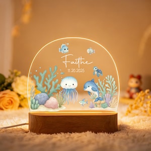 May include: A personalized night light featuring an underwater scene with a jellyfish, dolphin, and fish. The acrylic panel is illuminated by a wooden base, with the name "Faithe" and a date printed on it. The design includes coral and rocks.