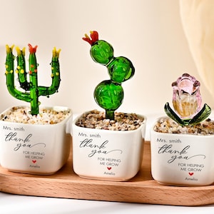 Könnte beinhalten: Drei Glaspflanzenfiguren in weißen, quadratischen Keramiktöpfen, die mit kleinen Kieselsteinen gefüllt sind. Die Töpfe tragen den aufgedruckten Text: Mrs. Smith, thank you for helping me grow, und Amelia. Die Pflanzen umfassen zwei grüne Kakteen und eine rosa Tulpe.