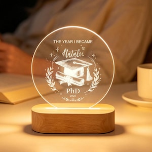 Könnte beinhalten: Ein rundes Acryl-Nachtlicht mit Holzsockel, graviert mit einer Doktorhut-Abbildung, einem Diplom und dem Text "THE YEAR I BECAME Natalie PhD 2023", umrahmt von einem Lorbeerkranz-Design, das ein warmes Licht auf den Tisch wirft.