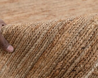 May include: Close-up of a natural fiber rug with a braided texture. The rug is a warm, earthy brown color, with variations in tone. The edge of the rug is being held up, revealing the woven construction.