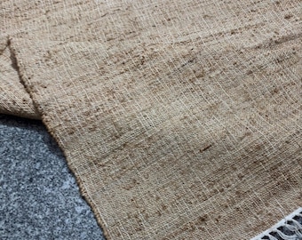 May include: A rectangular, woven jute rug in shades of tan and brown. The rug has a textured surface and a fringe detail along one edge. The natural fibers create a rustic, neutral aesthetic, suitable for various interior styles.