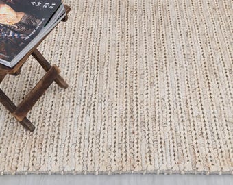 May include: A cream-coloured woven jute rug with a vertical texture. A small, dark brown wooden side table holds a magazine with a colourful cover. The rug's dimensions are not visible.