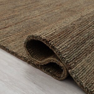 May include: A rolled-up brown jute rug with a textured surface. The rug's fibres are tightly woven, creating a durable surface. The edges of the rug are visible, showing the thickness and construction of the material. The colour palette is neutral, with shades of brown and tan.