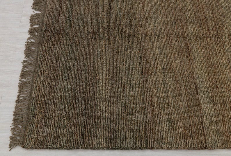 May include: Close-up of a brown woven rug with a fringed edge. The rug has a textured surface with vertical lines and varying shades of brown. The rug is made of natural fibres.