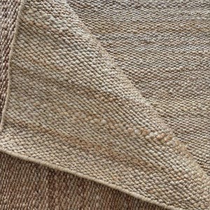 Op de afbeelding: Close-up van een natuurvezel tapijt met een geweven textuur. Het tapijt is lichtbruin van kleur met een opgevouwen hoek, waardoor de onderkant zichtbaar is. De weving is strak en uniform, waardoor een duurzaam en gestructureerd oppervlak ontstaat. Ideaal voor interieurdecoratie.
