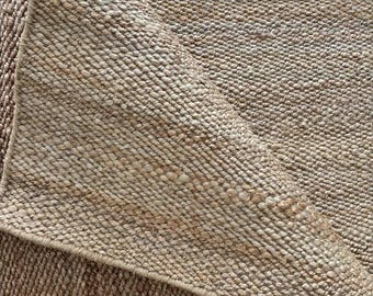 May include: Close-up of a natural fibre rug with a woven texture. The rug is a light tan colour with a folded corner, revealing the underside. The weave is tight and uniform, creating a durable and textured surface. Ideal for home decor.