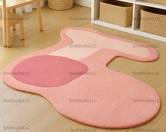 May include: A pink, abstract-shaped rug with varying shades of pink. The rug has a soft, plush texture and features a large, circular design in the center. The rug is placed on a light-colored wooden floor.