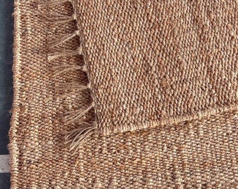 May include: Close-up of a natural fiber rug with a woven texture. The rug is a warm tan color, with a fringe detail along one edge. The rug is made of a durable material, suitable for high-traffic areas. The rug is a neutral color, making it versatile for any decor.