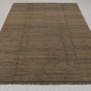 May include: A rectangular brown rug with a textured surface and a fringe along the bottom edge. The rug has a simple, neutral design, suitable for various interior styles. The rug is made of a woven material.