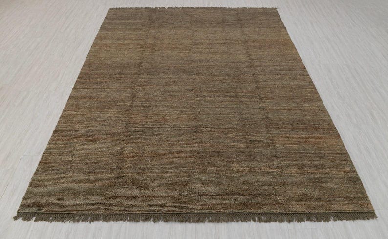 May include: A rectangular brown rug with a textured surface and a fringe along the bottom edge. The rug has a simple, neutral design, suitable for various interior styles. The rug is made of a woven material.
