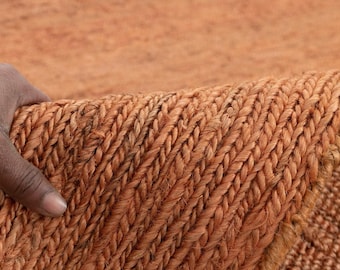 May include: Close-up of a woven rug in shades of orange and brown. The rug has a thick, textured appearance with a braided pattern. The rug is being held up, revealing the underside and the detailed weave.