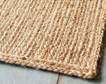 Puede incluir: Primer plano de una alfombra rectangular de yute tejido. La alfombra tiene un color marrón claro natural con un borde trenzado. La textura es visible, mostrando las fibras individuales y el patrón de tejido diagonal.