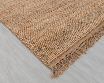May include: A close-up of a rectangular, woven jute rug. The rug is a light brown colour with a textured surface and a fringed edge. The rug is placed on a light grey surface.
