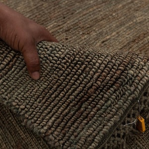 Puede incluir: Primer plano de una alfombra tejida con superficie texturizada. La alfombra es principalmente marrón y verde con un detalle de flecos. La alfombra se levanta, revelando su reverso. Una etiqueta amarilla está adherida a la alfombra.