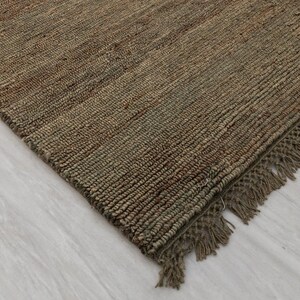 May include: A close-up shot of a brown jute rug with a woven texture and fringed edges. The rug has a natural, earthy colour and is placed on a light-coloured surface. The rug's design is simple and rustic.