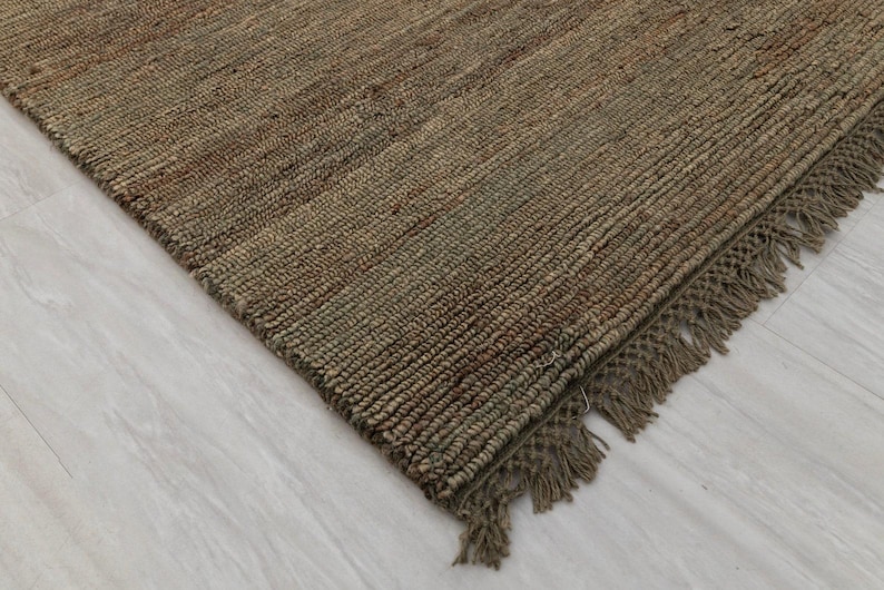 May include: A close-up shot of a brown jute rug with a woven texture and fringed edges. The rug has a natural, earthy colour and is placed on a light-coloured surface. The rug's design is simple and rustic.