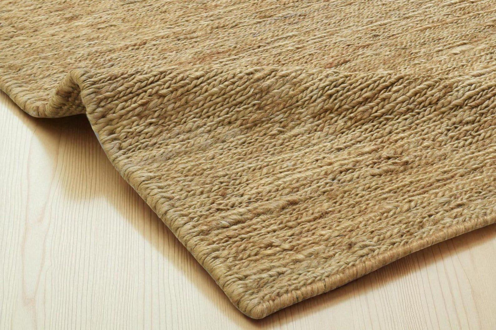 Könnte beinhalten: Nahaufnahme eines Naturfaserteppichs mit gewebter Textur. Der Teppich hat eine hellbraune Farbe und eine aufgerollte Kante, die die Unterseite freilegt. Der Teppich liegt auf einem hellen Holzboden.
