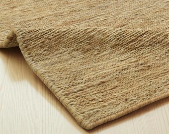 May include: A close-up of a natural fiber rug with a woven texture. The rug is a light tan color and has a rolled-up edge, revealing the underside. The rug is placed on a light-colored wooden floor.