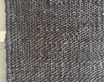 May include: A close-up of a dark gray woven rug with a natural fiber border. The rug has a textured surface with a diagonal pattern. The border is a light tan color.