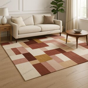 Könnte beinhalten: Ein rechteckiger Teppich mit geometrischem Muster in Rot-, Rosa-, Beige- und Goldtönen. Der Teppich liegt auf einem Holzboden. Ein beiges Sofa, ein runder Couchtisch aus Holz und ein Beistelltisch mit einer Lampe befinden sich im Hintergrund.