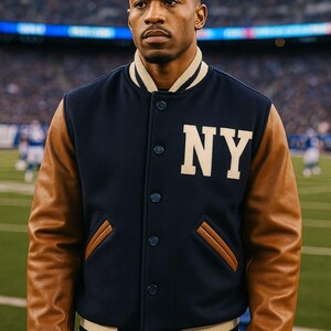 Può includere: Una giacca college blu navy con maniche in pelle marrone chiaro. La giacca presenta le lettere "NY" in bianco sul petto e ha colletto e polsini a righe. La giacca è abbottonata e indossata da una persona su un campo da calcio.