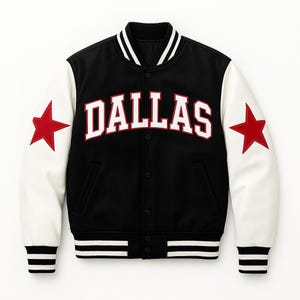 Op de afbeelding: Een zwart-witte varsity-jas met het woord "DALLAS" in wit met een rode omlijning over de borst. De jas heeft witte mouwen met rode sterpatches en zwart-wit gestreepte manchetten en zoom.
