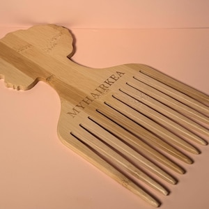 May include: A light brown wooden hair pick with a woman's head silhouette and the text "MYHAIRKEA". The pick has long, evenly spaced teeth, designed for styling and detangling hair. Made from wood.