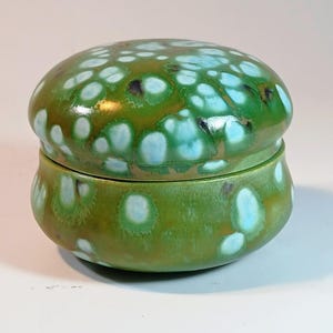 May include: A round ceramic lidded jar featuring a glossy finish with a mottled green and brown base color. The surface is decorated with distinct, circular crystalline patterns in shades of light blue and white, creating a spotted effect.