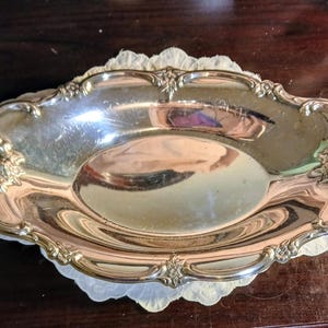 May include: A silver-colored, oval-shaped serving tray with ornate floral detailing around the edges. The tray sits on a white, scalloped doily. The reflective surface of the tray shows a distorted reflection.