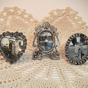 May include: Three silver-toned metal picture frames. The left frame is heart-shaped with a textured border. The center frame is oval with an ornate floral and leaf design. The right frame is oval with a border of small raised heart shapes.