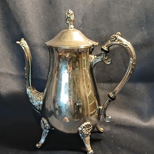 May include: Silver-colored coffee pot with a lid and ornate handle. The pot has a curved spout and sits on three decorative feet. The handle has a black accent. The pot is reflective, showing the surrounding environment.