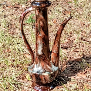 May include: A tall, brown ceramic pitcher with a long spout and curved handle. The pitcher has a marbled effect with shades of brown, red, and a hint of blue. It sits on a round base and is displayed outdoors on grass.