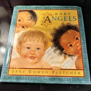 May include: A children's book titled "Baby Angels" features illustrations of three babies with angel wings. The cover has a gold border and the author's name, Jane Cowen-Fletcher, is printed at the bottom.