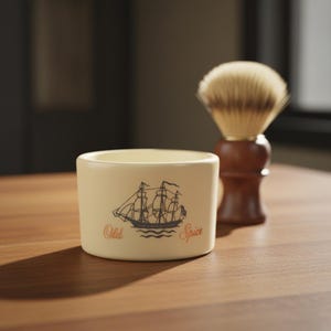 May include: A cream-colored shaving bowl with the words "Old Spice" and a ship illustration. A wooden-handled shaving brush with a light brown bristle head is behind the bowl. The items are on a wooden surface.