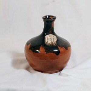 May include: A handmade ceramic bottle with a cork stopper. The bottle has a rounded base and a narrow neck. The glaze is a mix of brown and black, with a glossy finish. The cork stopper is visible on the side of the bottle.