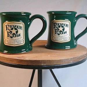 May include: Two dark green ceramic mugs with large handles. Each mug features a cream-colored label with the words "River Run Bed & Breakfast Inn" and a star graphic. The text "Kerrville, Texas" is also visible on the label.