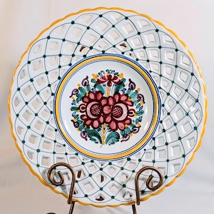 May include: White ceramic decorative plate with a lattice design and a floral center. The plate features a yellow scalloped edge and a central design with red, green, and blue floral elements. The plate is displayed on a decorative metal stand.