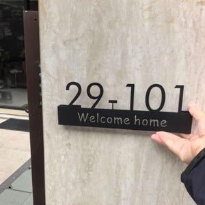 以下が含まれることがあります： 黒い金属製の家の番号標識で、「29-101」の数字と「Welcome home」の文字がサンセリフ体で表示されています。標識は明るい色のテクスチャのある壁に取り付けられています。