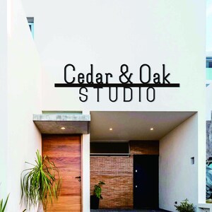 以下が含まれることがあります： 木製のドアと、入り口の上に黒い文字で「Cedar & Oak STUDIO」と書かれたモダンな建物の外観。建物は白いファサードとコンクリートのオーバーハングがあります。