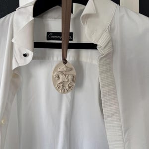 May include: A white shirt with a decorative oval pendant hanging from a brown ribbon. The pendant features a relief design of a horse-drawn carriage. The shirt is hanging on a black hanger.