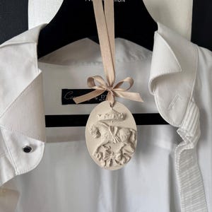 Puede incluir: Una camisa blanca cuelga de una percha negra, con un adorno decorativo de forma ovalada suspendido de una cinta. El adorno presenta un diseño en relieve detallado de un carrusel con caballos y una corona.