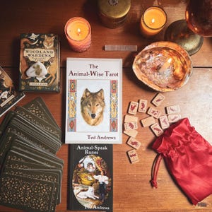 May include: A collection of spiritual items on a wooden surface. Includes tarot decks, books titled "The Animal-Wise Tarot" and "Animal-Speak Runes", wooden runes, candles, and a red drawstring bag. The book covers feature animal illustrations.