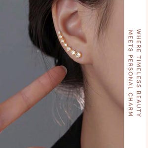 Może przedstawiać: Złoty kolczyk typu ear climber z serią stopniowanych, przezroczystych kryształów. Kolczyk zakrzywia się wzdłuż ucha, tworząc błyszczący akcent. Obraz zawiera również tekst "Gemoura Jewels" i "Where Timeless Beauty Meets Personal Charm."