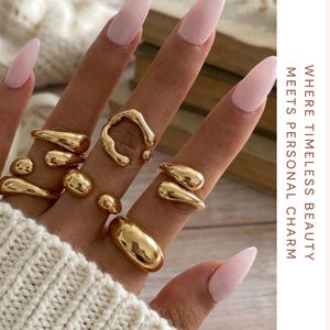 Könnte beinhalten: Nahaufnahme einer Hand mit mehreren goldfarbenen Ringen in einzigartigen, organischen Formen. Die Ringe sind glatt und glänzend. Der Text "GemouraJewels" und "Where timeless beauty meets personal charm" ist ebenfalls enthalten.