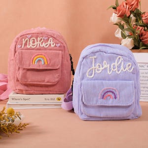 Könnte beinhalten: Zwei kleine Rucksäcke aus Cord, einer rosa und einer lila, mit gestickten Namen und Regenbogen-Designs. Der rosa Rucksack trägt den Namen "Nora", der lila Rucksack "Jordie". Beide haben eine kleine Vordertasche.